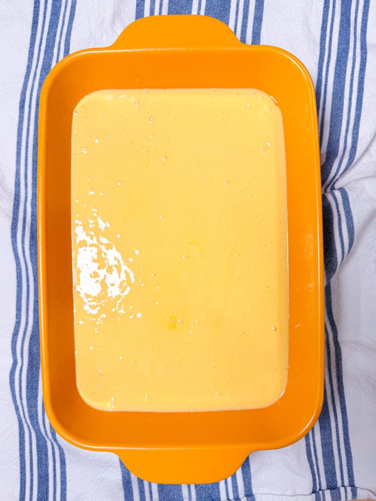 An orange rectangular baking dish filled with a smooth yellow batter sits on a white towel with blue stripes.