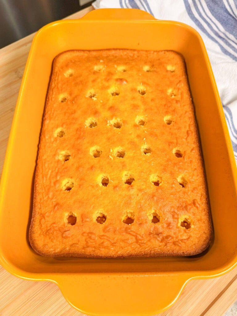A rectangular yellow baking dish holds a golden-brown sheet cake with evenly spaced holes on top, sitting on a wooden surface with a striped cloth in the background.