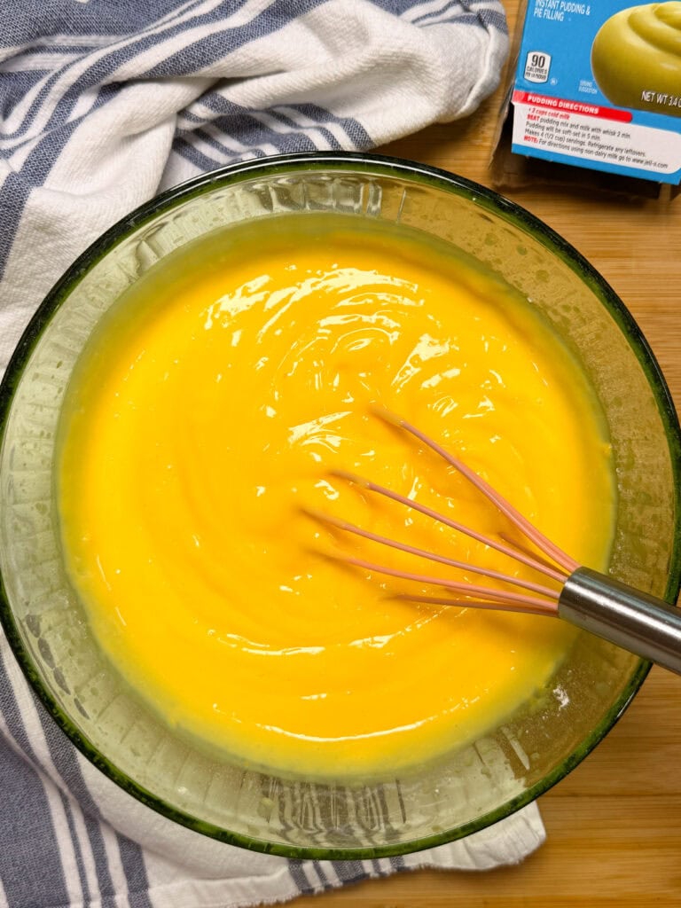 A glass bowl filled with smooth yellow pudding is being whisked. A blue and white striped towel and part of a boxed pudding mix are visible nearby on a wooden surface.