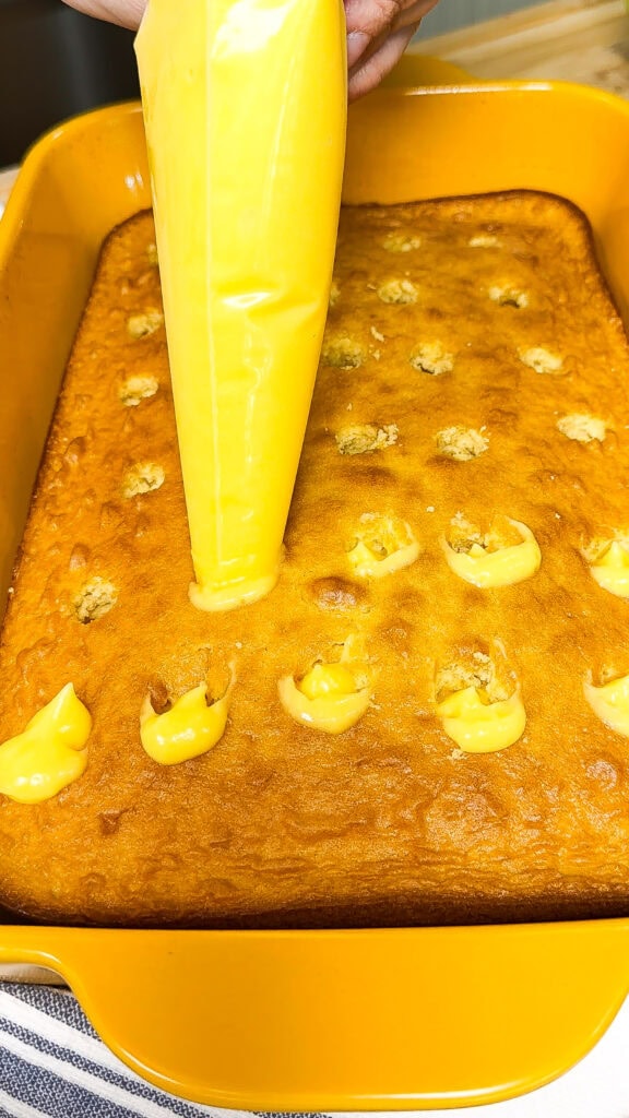 A hand uses a piping bag to fill holes in a golden sheet cake with yellow cream inside a yellow baking dish.
