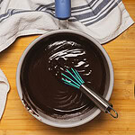 A saucepan filled with glossy melted chocolate and a whisk sits on a wooden surface, next to a white towel with blue stripes.