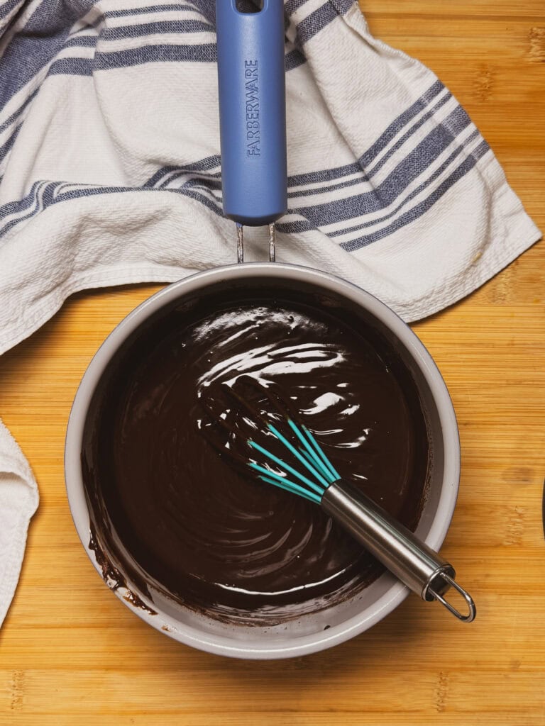 A saucepan filled with glossy melted chocolate and a whisk sits on a wooden surface, next to a white towel with blue stripes.