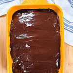 A yellow baking dish filled with a dessert topped with a smooth, glossy layer of chocolate, sitting on a wooden surface with a white and blue towel nearby.