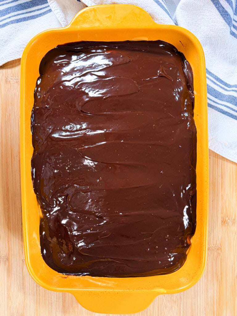 A yellow baking dish filled with a dessert topped with a smooth, glossy layer of chocolate, sitting on a wooden surface with a white and blue towel nearby.