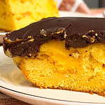 A close-up of a slice of yellow cake with a chocolate frosting layer, showing a moist texture and a visible filling, on a white plate. Another slice is blurred in the background.