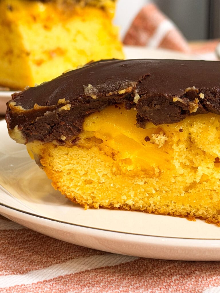 A close-up of a slice of yellow cake with a chocolate frosting layer, showing a moist texture and a visible filling, on a white plate. Another slice is blurred in the background.