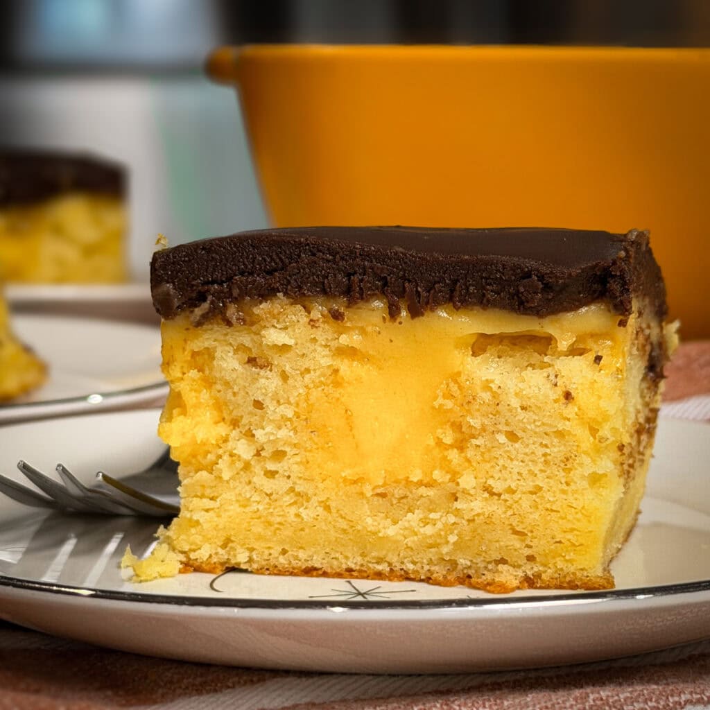 A slice of yellow cake with a creamy filling and a thick layer of chocolate frosting sits on a plate next to a fork, with a blurred orange bowl in the background.