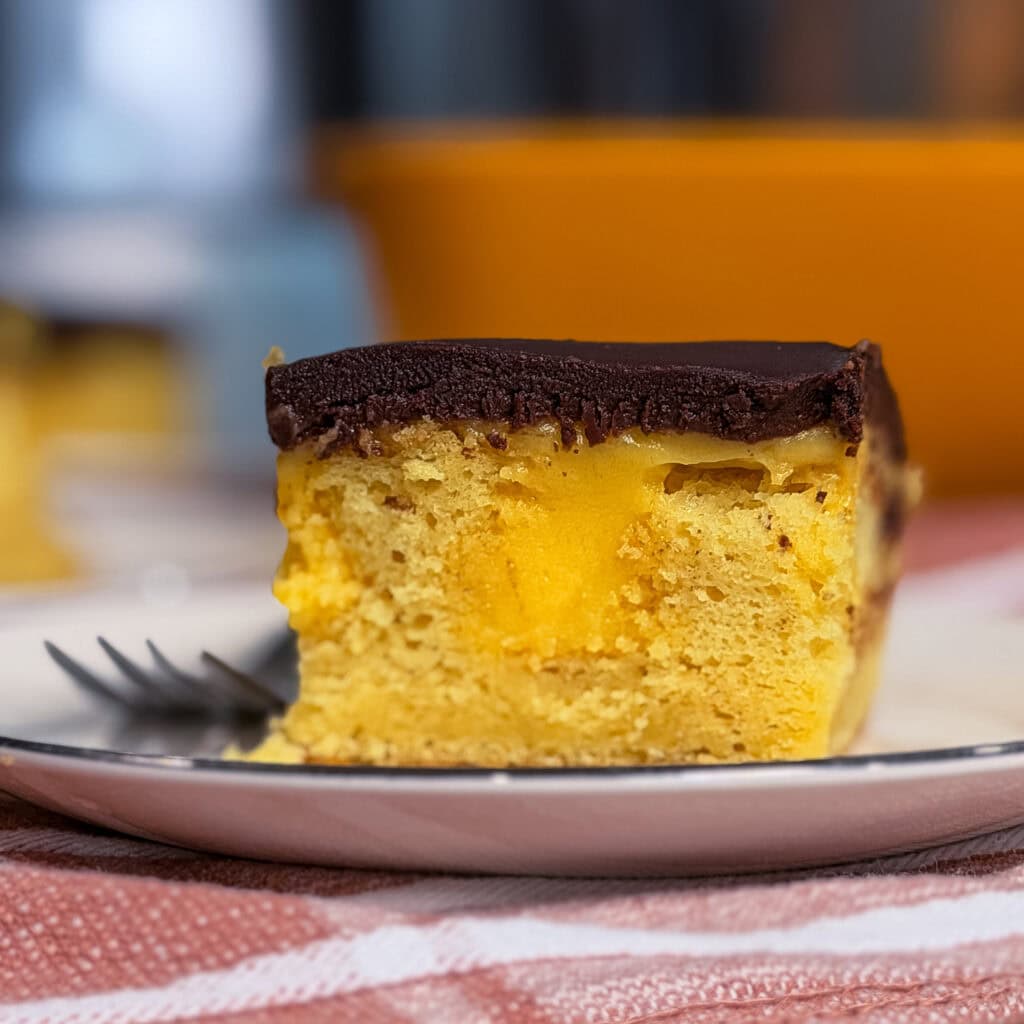 A close-up of a slice of yellow cake with chocolate icing and a custard or cream filling, served on a white plate with a fork beside it. The background is softly blurred.