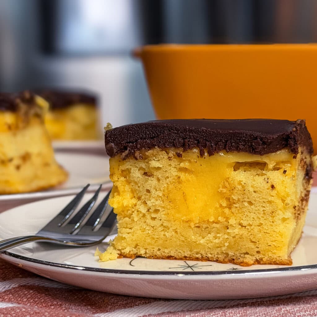 A slice of yellow sponge cake with creamy filling and a chocolate topping sits on a plate with a fork. A blurred yellow bowl and another cake slice are in the background.