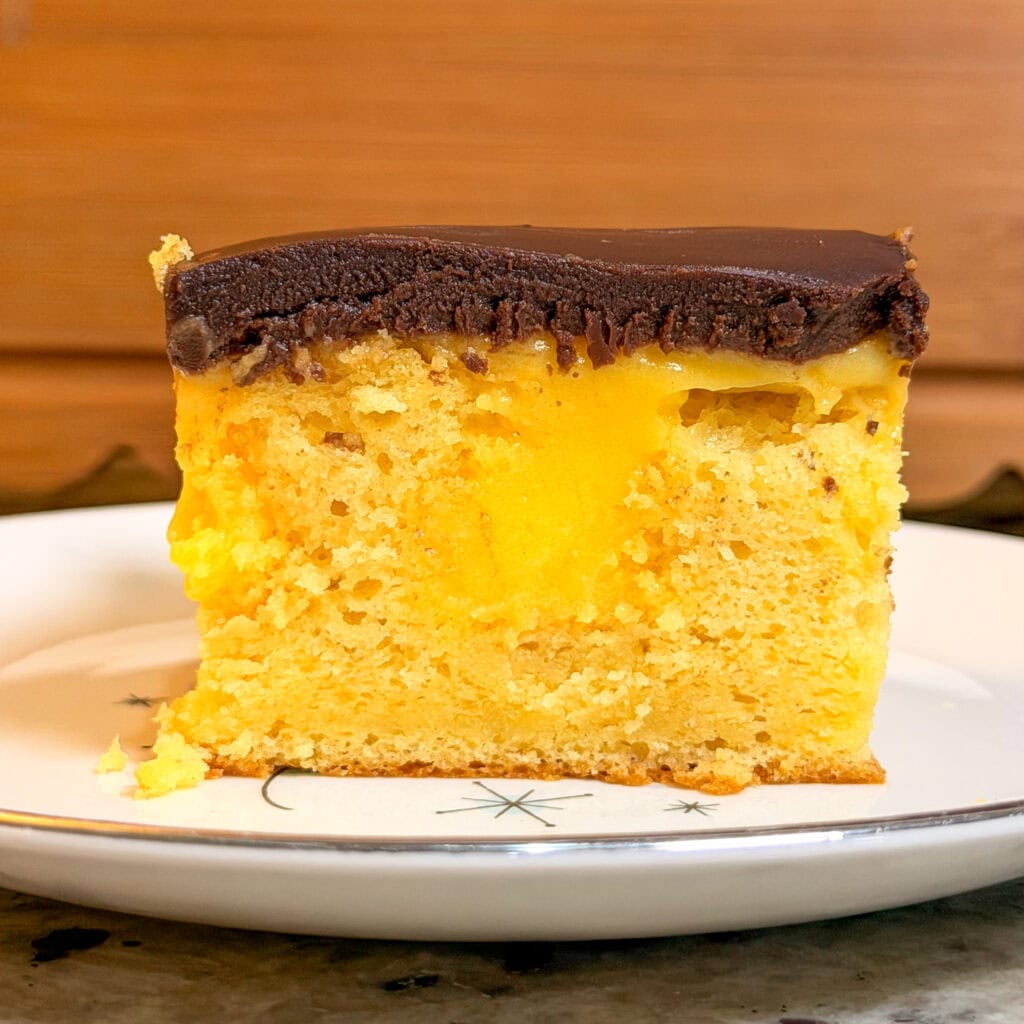 A slice of yellow cake with a gooey yellow filling and a thick layer of chocolate frosting on top, served on a white plate with star designs.