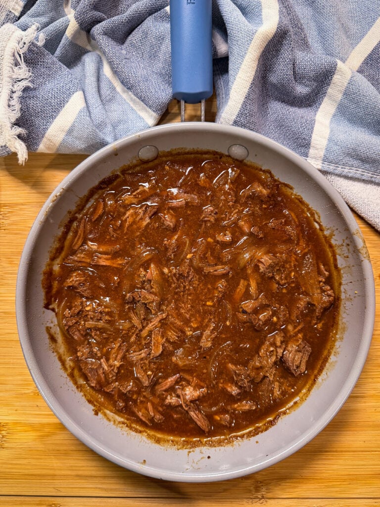 A skillet filled with shredded beef in a rich brown sauce sits on a wooden surface, with a blue and white striped towel partially visible at the top.