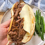 A hand holds a sandwich filled with shredded beef and a slice of cheese on a bun, above a plate with green beans and a blue-and-white towel nearby.