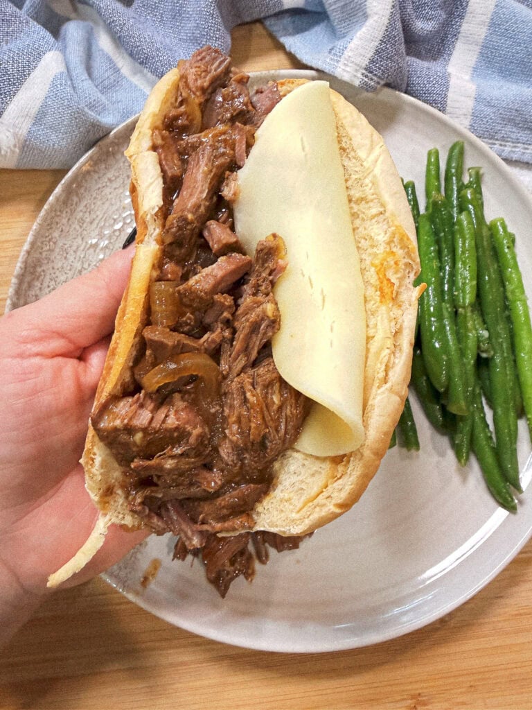 A hand holds a sandwich filled with shredded beef and a slice of cheese on a bun, above a plate with green beans and a blue-and-white towel nearby.