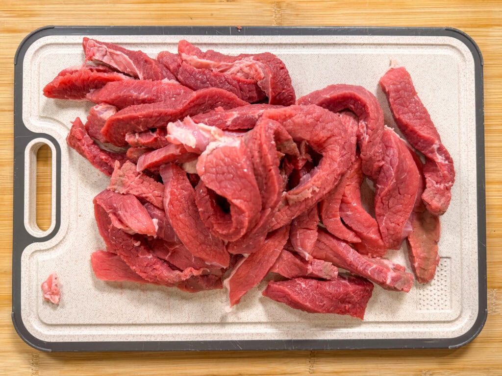 Sliced raw beef strips are spread out on a white cutting board with a black and white border, placed on a wooden surface.