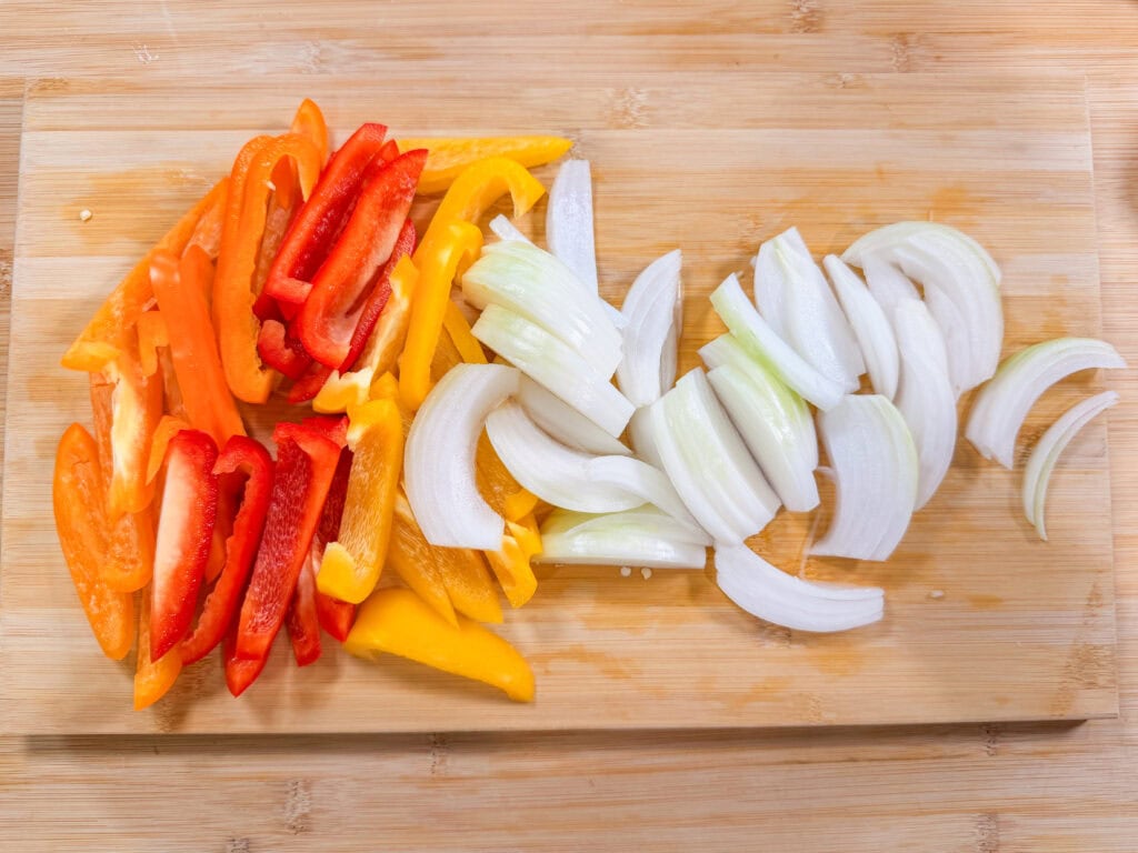Sliced red, orange, and yellow bell peppers and white onions arranged on a wooden cutting board.
