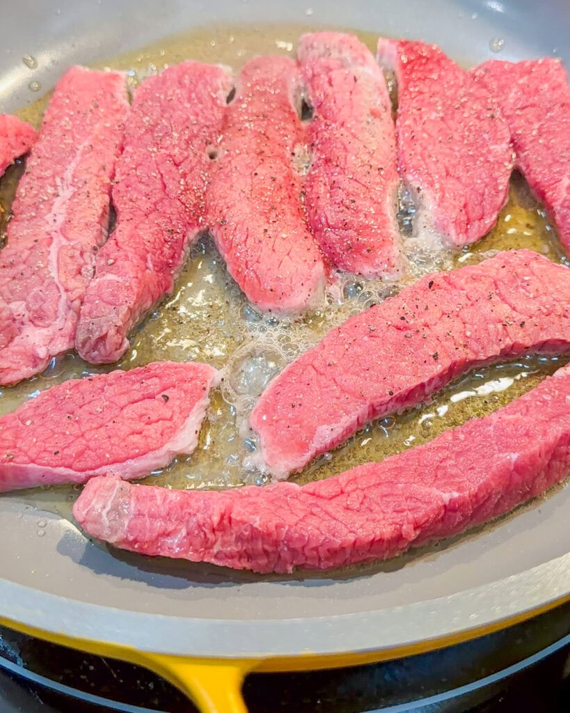 Several strips of raw beef are being cooked in a pan with bubbling oil. The meat is seasoned with black pepper and is starting to brown at the edges.