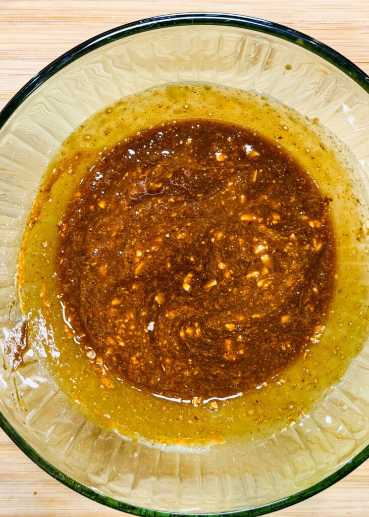A glass bowl containing a brown, oily marinade with visible minced garlic and spices sits on a light wooden surface.