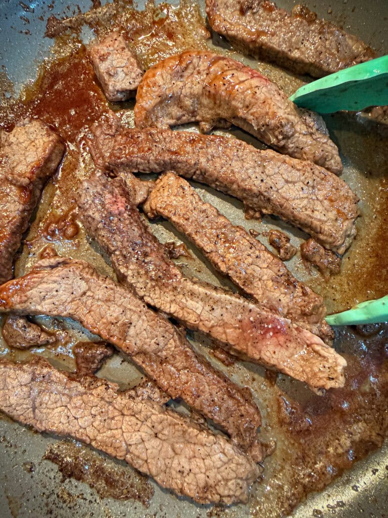 Strips of beef sizzling in a pan, with green tongs holding one piece. The meat appears browned and juicy, with visible seasoning and a bit of liquid pooling in the pan.