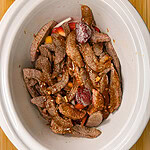 Raw beef strips and chopped vegetables with sauce in a white slow cooker, placed on a wooden surface.