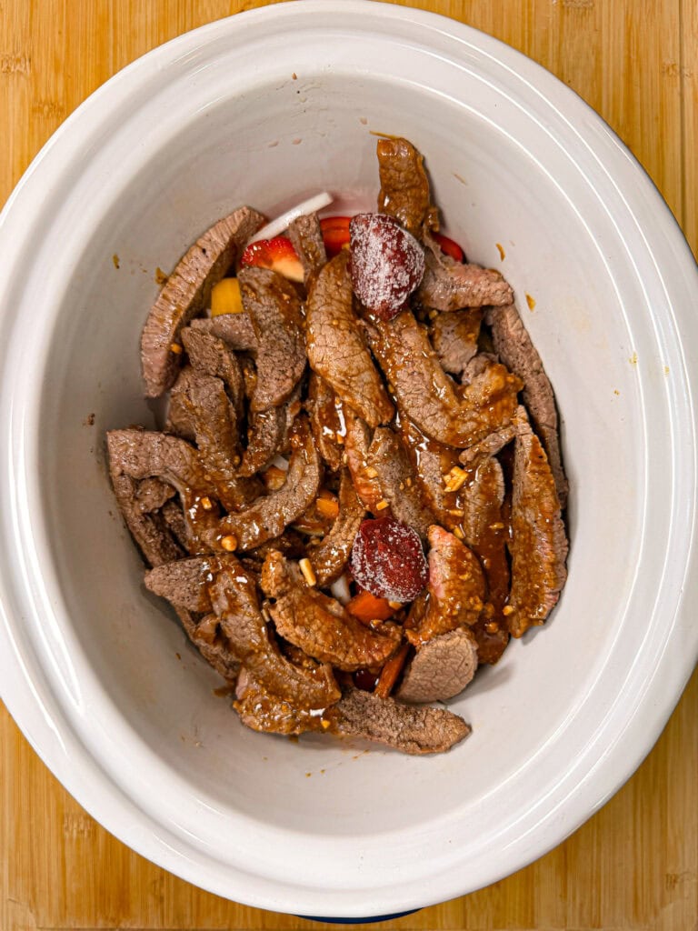 Raw beef strips and chopped vegetables with sauce in a white slow cooker, placed on a wooden surface.