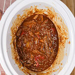 A white slow cooker filled with beef stew in a rich brown sauce, visible slices of beef, peppers, onions, and sesame seeds, on a wooden surface next to a striped towel.
