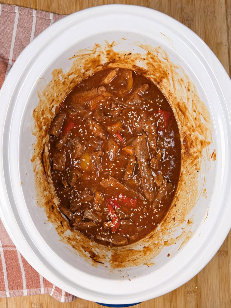 A white slow cooker filled with beef stew in a rich brown sauce, visible slices of beef, peppers, onions, and sesame seeds, on a wooden surface next to a striped towel.