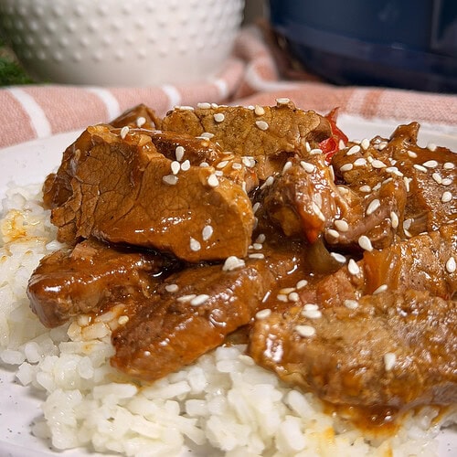 Slices of beef in a brown sauce with sesame seeds, served over white rice on a plate. In the background, a bowl with colorful vegetable sticks and a blue object are visible.