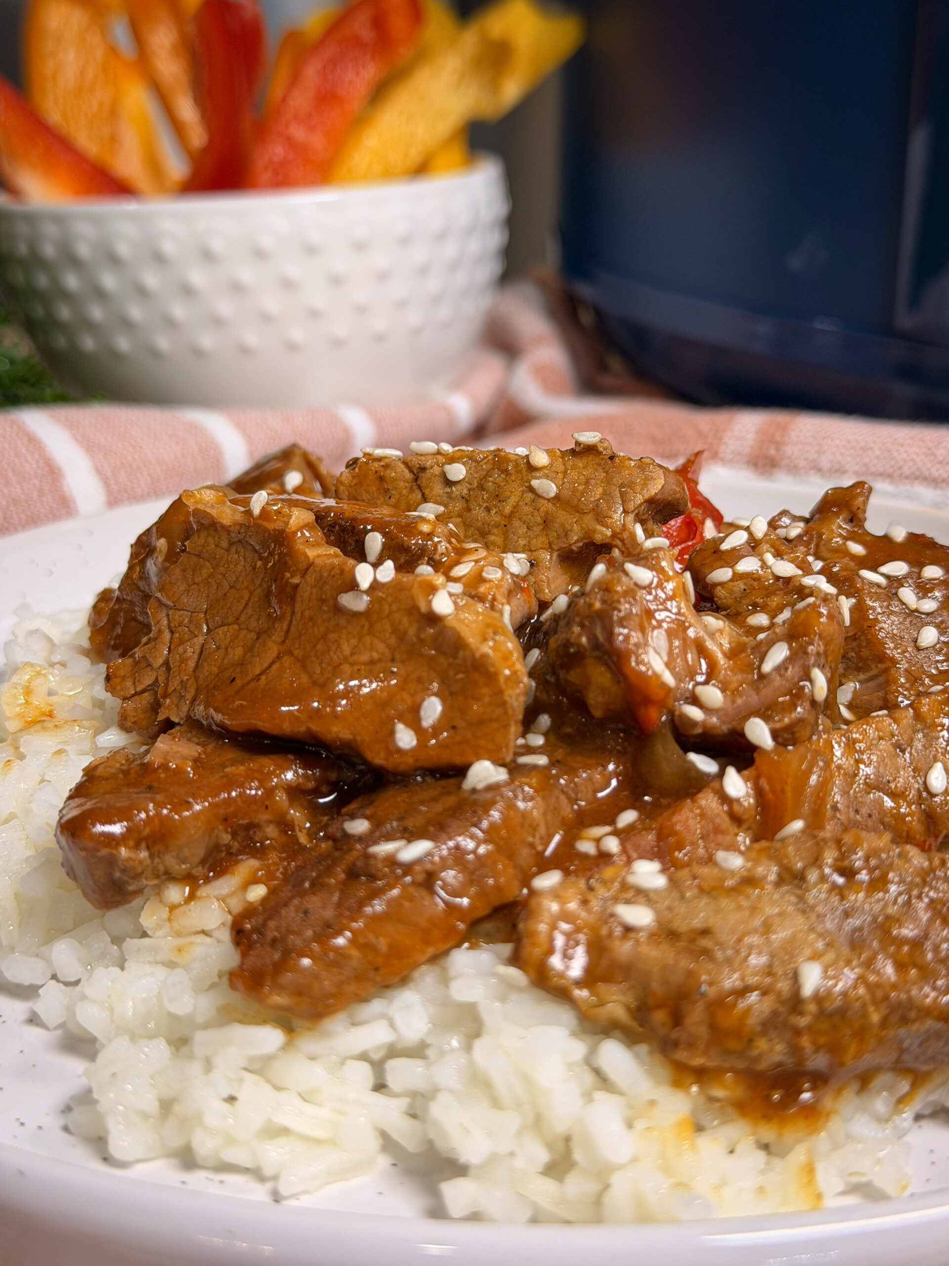 Plate of white rice topped with saucy beef slices and sesame seeds, with a bowl of colorful bell pepper strips in the background and a striped cloth underneath.