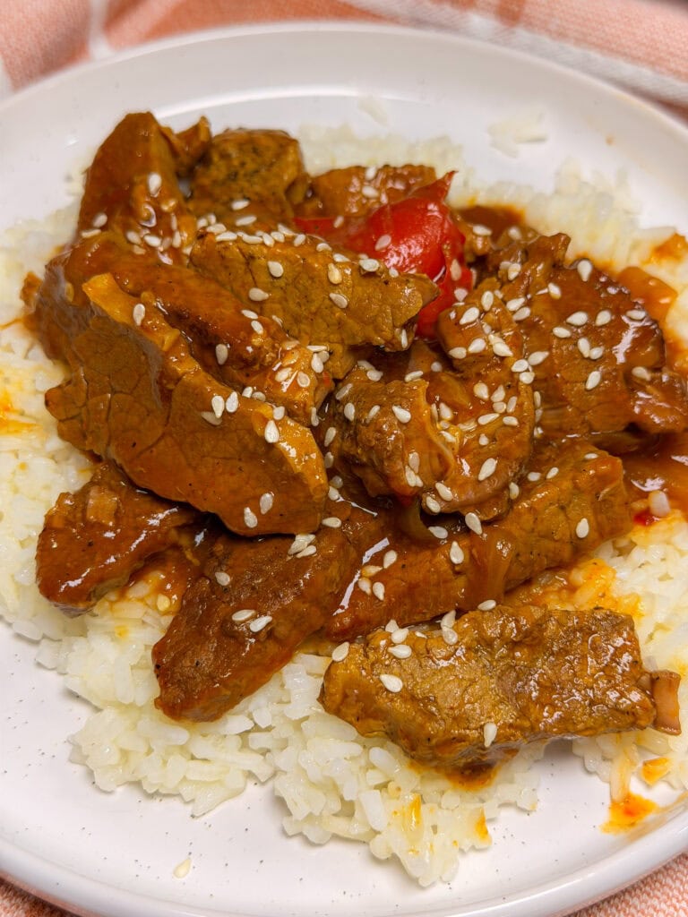 A plate of white rice topped with saucy beef strips, red bell peppers, and sprinkled with white sesame seeds.