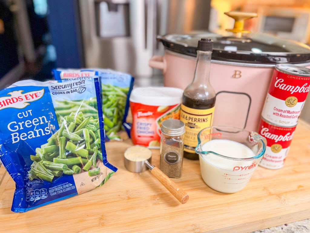 Ingredients for a casserole are on a wooden board, including green beans, milk, cream of mushroom soup, soy sauce, pepper, cheese, and a slow cooker in the background.