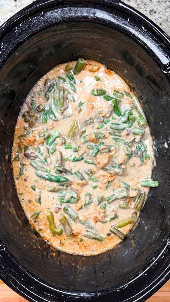 Green bean casserole with creamy sauce and mushrooms in a black slow cooker, shown from above on a kitchen countertop. The mixture is cooked, with visible chunks of green beans and sauce.
