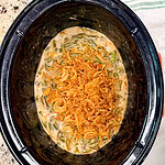 A slow cooker filled with green bean casserole topped with crispy fried onions, sitting on a speckled countertop next to a folded green and white striped kitchen towel.