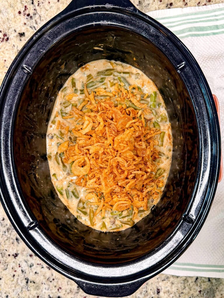 A slow cooker filled with green bean casserole topped with crispy fried onions, sitting on a speckled countertop next to a folded green and white striped kitchen towel.