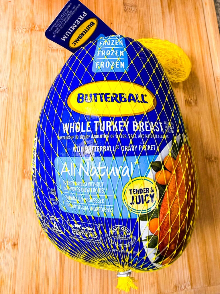 A packaged Butterball frozen whole turkey breast, wrapped in yellow netting, sits on a wooden surface. The label highlights All Natural and Tender & Juicy..