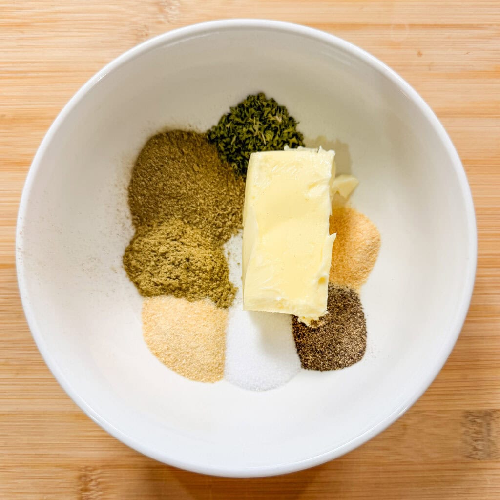 A white bowl containing a pat of butter surrounded by small piles of various ground spices and herbs sits on a wooden surface.
