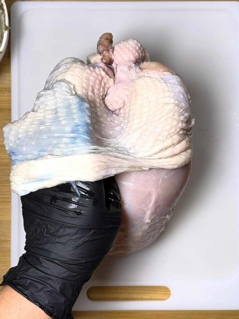 A hand wearing a black glove is lifting the skin of a raw chicken breast, exposing the flesh underneath, on a white cutting board.