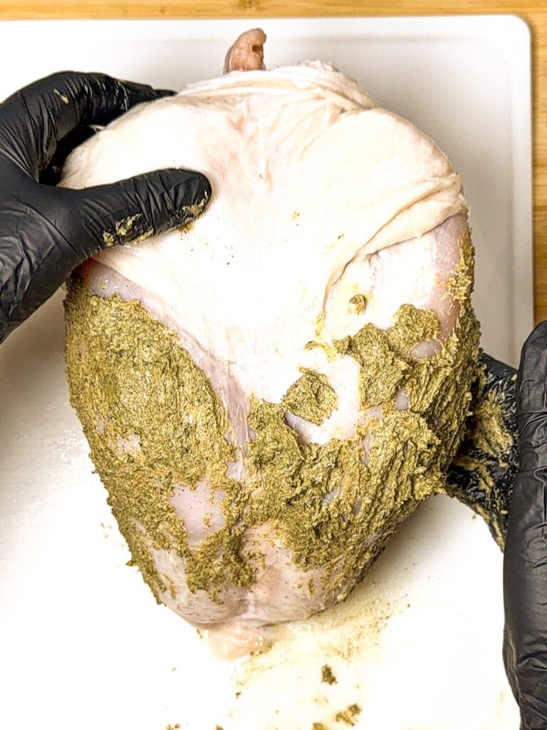 Gloved hands rub a green herb paste onto a raw turkey breast sitting on a white cutting board.