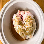 Raw, seasoned turkey breast in a white oval slow cooker, sitting on a wooden surface. The turkey is coated with spices and ready to be cooked.