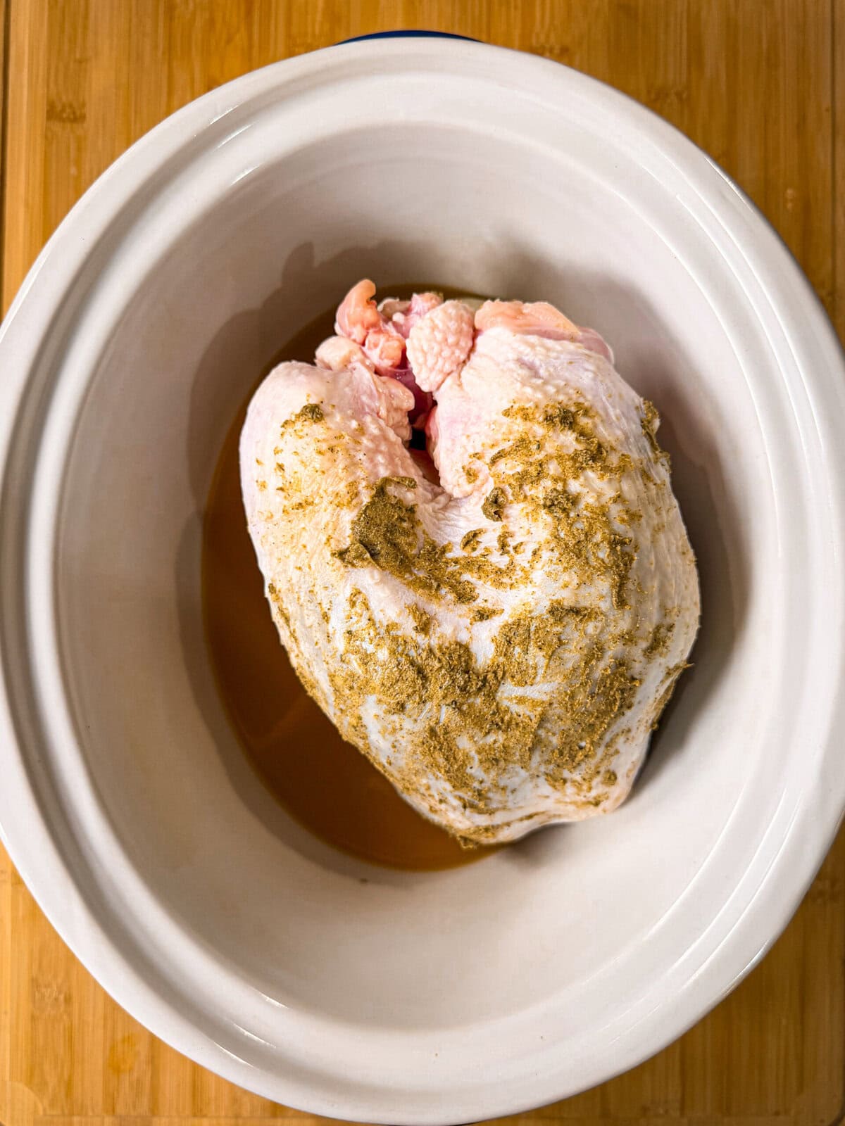 Raw, seasoned turkey breast in a white oval slow cooker, sitting on a wooden surface. The turkey is coated with spices and ready to be cooked.