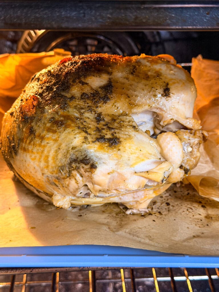 A seasoned whole turkey is roasting in an oven, sitting on parchment paper. The skin is golden brown, and the ovens heating element is visible in the background.