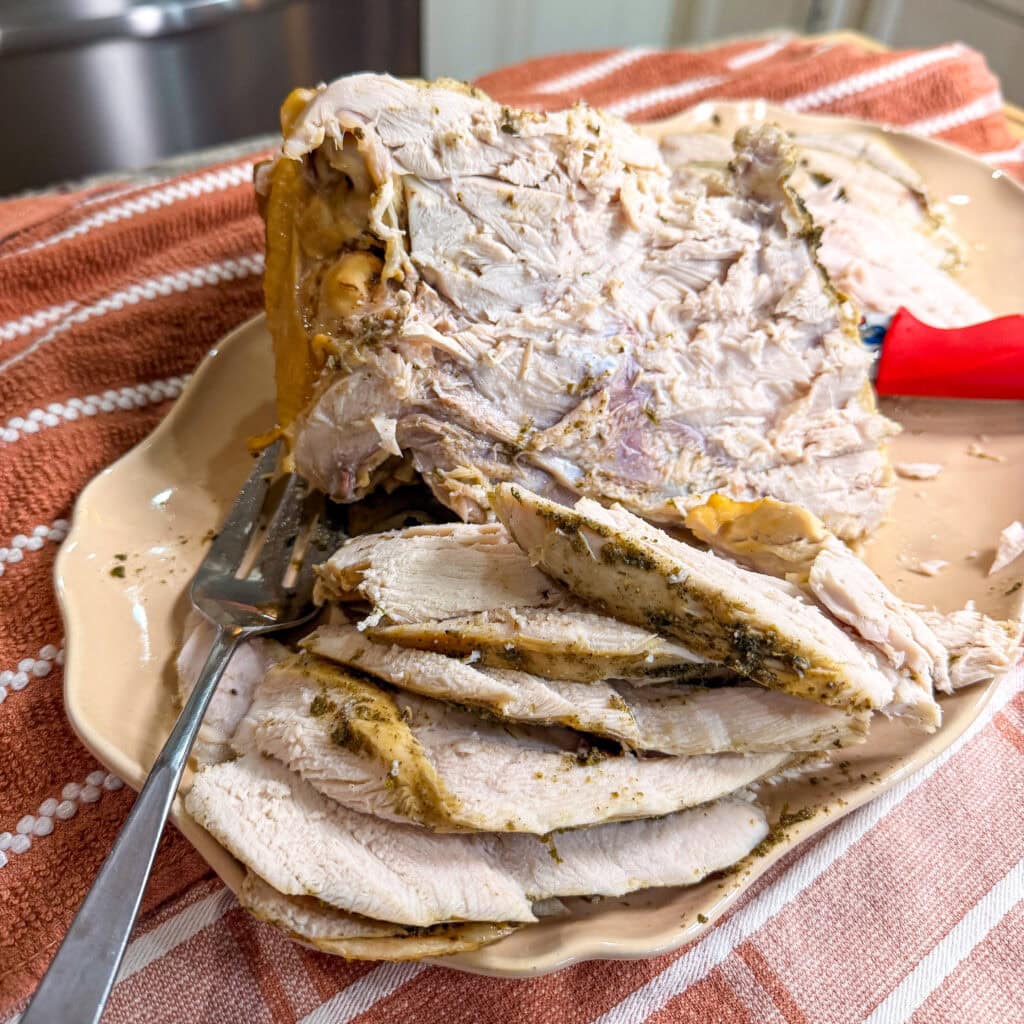 Sliced roasted turkey breast is arranged on a beige platter with a fork on the side. The platter sits on an orange and white striped towel.