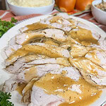 Sliced turkey with gravy on a white platter, served with mashed potatoes, bread rolls, and stuffing in the background on a kitchen counter.
