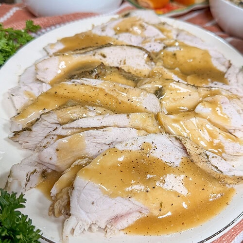 Sliced turkey with gravy on a white platter, served with mashed potatoes, bread rolls, and stuffing in the background on a kitchen counter.