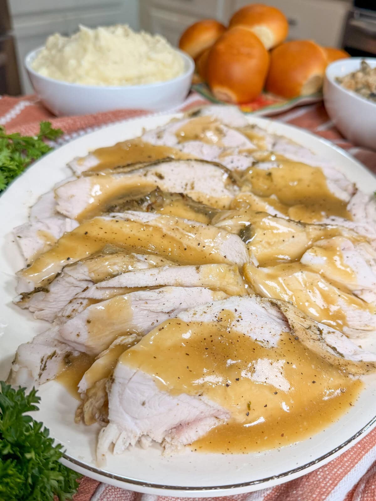 Sliced turkey with gravy on a white platter, served with mashed potatoes, bread rolls, and stuffing in the background on a kitchen counter.