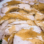 Sliced turkey breast covered in golden-brown gravy, arranged on a white plate, with visible seasoning and herbs on the meat and sauce.