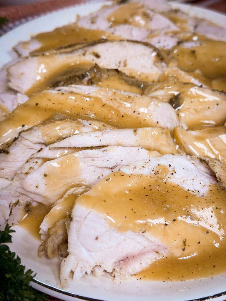 Sliced turkey breast covered in golden-brown gravy, arranged on a white plate, with visible seasoning and herbs on the meat and sauce.