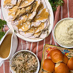 A Thanksgiving meal with sliced turkey and gravy, mashed potatoes, stuffing, green bean casserole, dinner rolls, a gravy boat, and parsley garnish on a striped cloth.