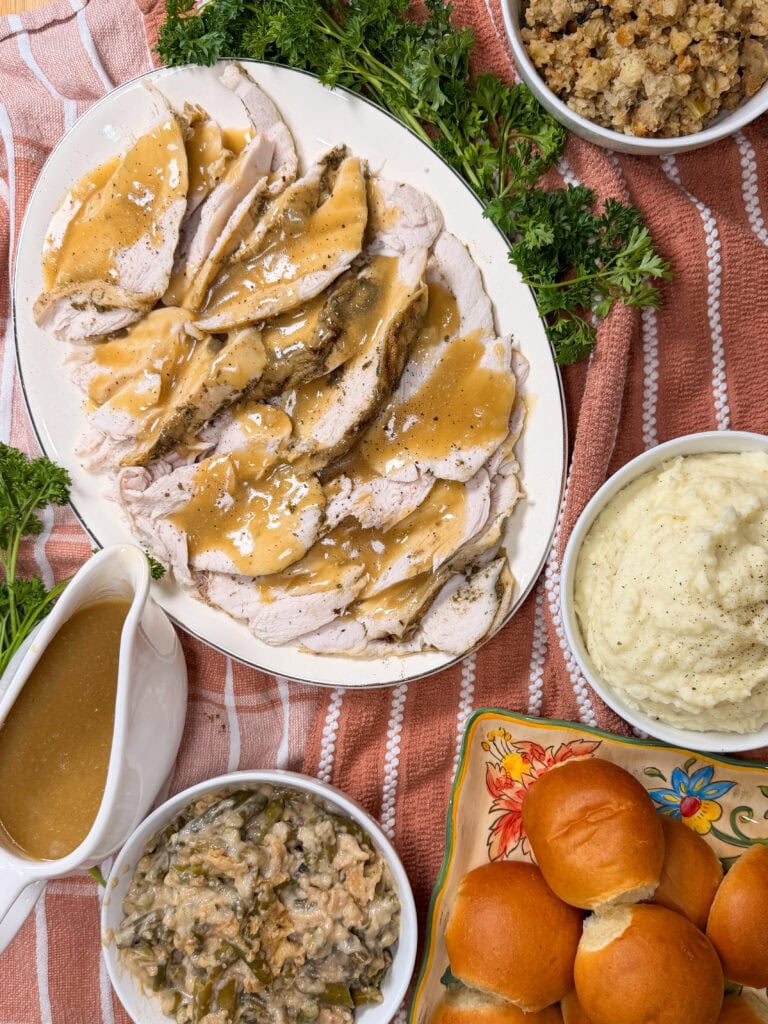 A Thanksgiving meal with sliced turkey and gravy, mashed potatoes, stuffing, green bean casserole, dinner rolls, a gravy boat, and parsley garnish on a striped cloth.