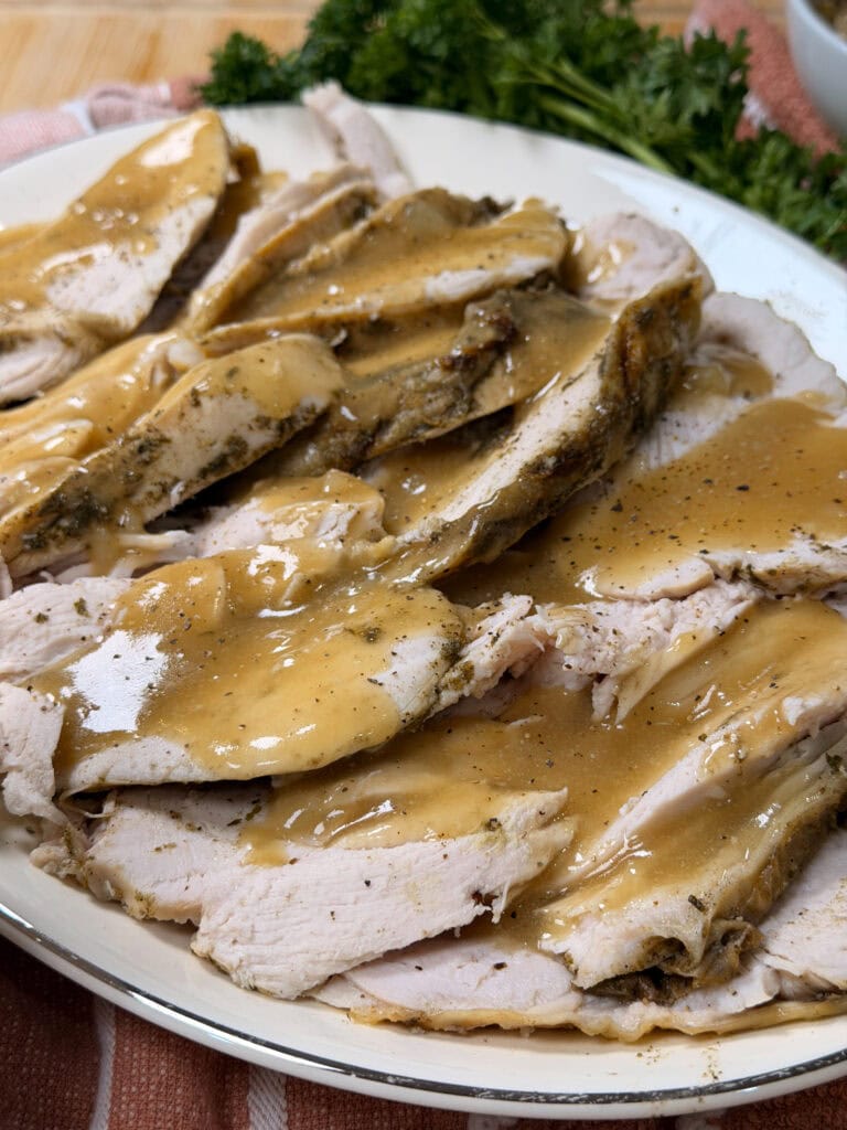 Slices of turkey breast topped with brown gravy are arranged on a white plate, with fresh parsley in the background and a striped cloth beneath the plate.