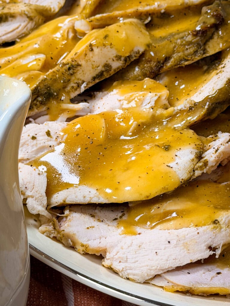 Close-up of sliced turkey breast topped with golden brown gravy on a plate, with part of a white gravy boat visible in the corner.